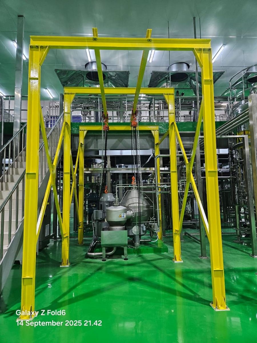 Rangka gantry baja cat kuning terpasang di atas peralatan produksi pada ruangan berlantai hijau